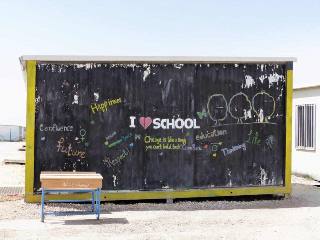 I love school A black-painted wall of a temporary school structure decorated with colorful, handwritten words and drawings. Phrases like 'I ❤️ SCHOOL,' 'Confidence,' 'Future,' 'Happiness,' 'Peace,' and a quote by Queen Rania are surrounded by childlike illustrations of trees, symbols, and hearts. A single wooden desk with blue legs is placed in front of the wall on a dirt ground under a clear sky. All rights reserved.