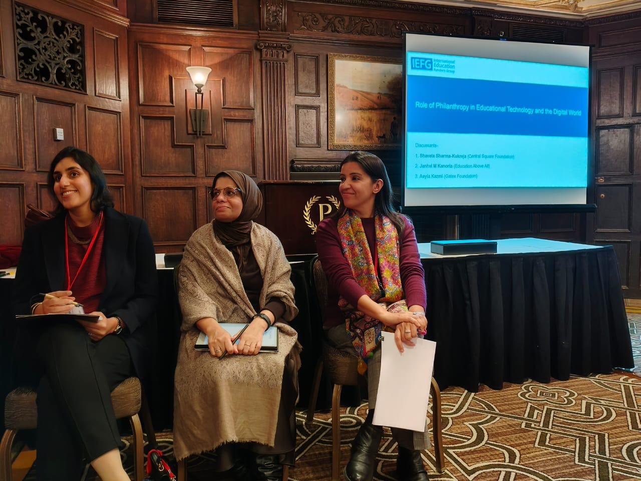 Three women seated in a panel discussion room with wood-paneled walls and a presentation screen showing 'Role of Philanthropy in Educational Technology and the Digital World'